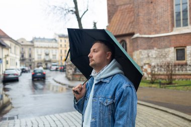 Kot ceketli ve kapüşonlu bir adam yağmurlu bir Avrupa caddesinde büyük siyah bir şemsiye tutarak yürüyor. Arka planda tarihi mimari ve ıslak kaldırım taşları yer alıyor, karamsar ve bulutlu bir görüntü yakalıyorlar.