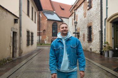 Kot ceketli ve kapüşonlu bir adam Avrupa 'nın tarihi bir şehrinde kaldırımlı bir caddede dururken gülümsüyor. Yağmurlu hava ve eski taş binalar büyüleyici ve huzurlu bir hava yaratıyor..