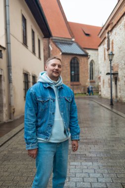 Kot ceketli ve kapüşonlu bir adam Avrupa 'nın tarihi bir şehrinde kaldırımlı bir caddede dururken gülümsüyor. Yağmurlu hava ve eski taş binalar büyüleyici ve huzurlu bir hava yaratıyor..