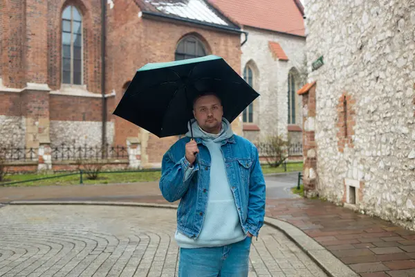 Kot ceketli ve kapüşonlu bir adam yağmurlu bir Avrupa caddesinde büyük siyah bir şemsiye tutarak yürüyor. Arka planda tarihi mimari ve ıslak kaldırım taşları yer alıyor, karamsar ve bulutlu bir görüntü yakalıyorlar.