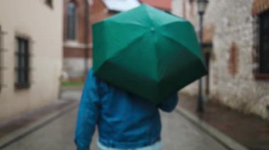 Avrupa 'nın tarihi bir şehrinde sakin, kaldırım taşı bir sokakta yürürken elinde yeşil bir şemsiye tutan bir adamın arka görüntüsü. Yağmurlu atmosfer ve eski binalar huzurlu ve karamsar bir ortam yaratıyor..