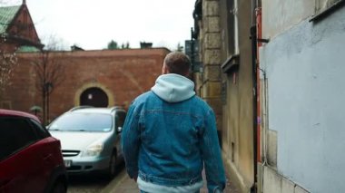 Kot ceketli ve kapüşonlu bir adam tarihi bir bölgede dar bir kaldırımda park edilmiş arabaların yanında yürüyor. Arka plandaki eski taş binalar ve tuğla duvarlar sonsuz bir şehir havası katıyor..