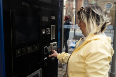 A woman in a yellow jacket uses a ticket machine in Krakow to buy a tram or bus ticket. The machine is located near a street with historical buildings in the background.