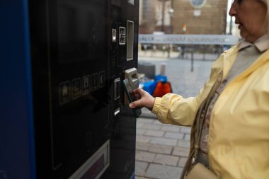 A woman in a yellow jacket uses a ticket machine in Krakow to buy a tram or bus ticket. The machine is located near a street with historical buildings in the background.