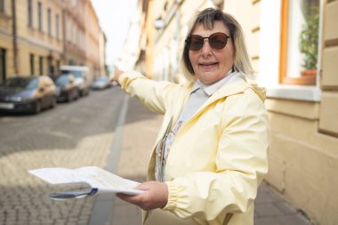 Smiling elderly woman in a yellow jacket and sunglasses holding a city map and pointing at something on a cobblestone street in a European town. She appears to be exploring or navigating her way. 