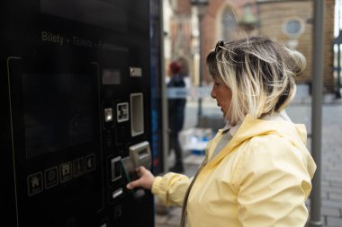 A woman in a yellow jacket uses a ticket machine in Krakow to buy a tram or bus ticket. The machine is located near a street with historical buildings in the background.