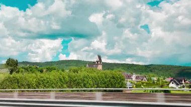 Dramatik bulutlu bir gökyüzünün altında ormanın üzerinde yükselen uzaktaki bir kilise kulesinin hareket halindeki bir aracının görüntüsü. Seyahat, tarih ve Avrupa kırsalını çağrıştıran huzurlu ve manzaralı bir an