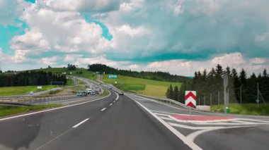 Yeşil tepeler, tarlalar ve ormanla çevrili çok şeritli bir otoyolda hareket halindeki bir arabanın içinden çekilen görüntüler. Bulutlu mavi gökyüzü dinamik seyahat sahnesini güçlendirir. Bunun gibi temalar için mükemmel