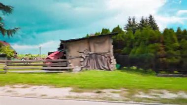 Hareket halindeki bir araçtan yüksek çam ağaçları, yeşil çimenler ve özel evleri olan sakin bir yerleşim bölgesini geçerken görüntülenmiş. Açık mavi gökyüzü altında huzurlu bir banliyö ya da kırsal manzara, temalar için ideal