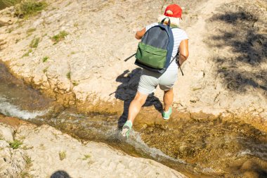 Kısa şortlu, kırmızı şapkalı ve sırt çantalı aktif bir son sınıf öğrencisi dağ yürüyüşü sırasında sığ bir dereyi geçiyor. Yukarıdan çekilen bu maceraperest an, hareketi, özgürlüğü ve sağlıklı bir açık havayı yakalar.