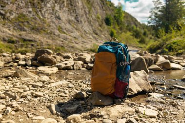 Mavi gökyüzünün altındaki manzaralı bir dağ vadisinde kayalık bir nehir yatağında dinlenen renkli sırt çantası. Macera sembolü, yürüyüş, solo seyahat ve doğayla bağlantı. Açık hava yaşam tarzı ve turizm için mükemmel.