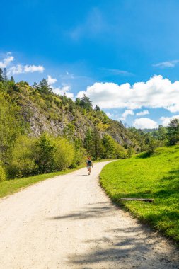 Mavi gökyüzünün altındaki yeşil bir dağ vadisinde çakıllı bir patikada bisiklet süren birinin dikey fotoğrafı. Ağaçlar, kayalıklar ve çimlerle çevrili açık hava aktivitesi için ideal bir yer.