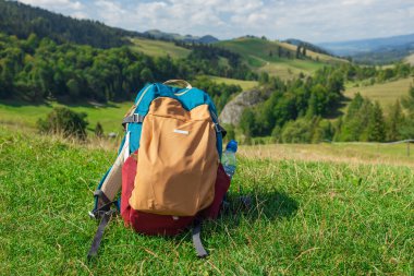 Renkli bir yürüyüş sırt çantası yemyeşil bir yamaçta uzanan manzaralı bir dağ vadisinde uzanıyor. Bu resim açık hava macerasını, özgürlüğü ve seyahat ruhunu sembolize ediyor. İdeal
