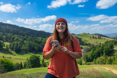 Sırt çantalı genç bir bayan gezgin güzel bir kırsal alana bakan çimenli bir patikada duruyor. Elinde bir harita ve akıllı telefon var. Tepelerde tek başına yürürken güzergahını kontrol ediyor.. 