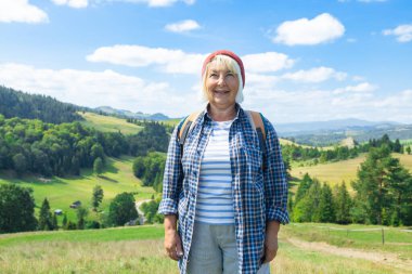 Yemyeşil bir yamaçta duran yaşlı bir kadın sırt çantasıyla, güzel panoramik dağ manzarasından etkilenmiş ve memnun görünüyor. Ekose gömlek ve kırmızı bereyle, aktif olarak temsil ediliyor.