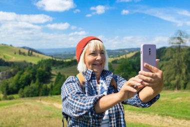 Gülümseyen son sınıf öğrencisi bir kadın, kayan tepelerin ve yemyeşil yeşilliklerin resimli manzarasında yürürken selfie çekiyor. Kırmızı bir bere ve ekose gömlek giyerek, doğanın özgürlüğünün ve