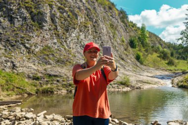 Yürüyüş kıyafetli, kırmızı şapkalı, kayanın üstünde oturmuş, huzurlu bir dağ deresinin yanında selfie çeken gülümseyen genç bir kadın. Doğal güzellik ve kayalık araziyle çevrelenmiş halde, ara verirken eğleniyor.