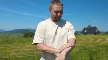 Genç bir adam manzaralı bir kırsal alanda duruyor ve parlak güneşin altında kollarına güneş kremi sürüyor. Görüntü yaz cilt bakımını, güneş korumasını ve yeşil ile açık hava yaşam tarzını yansıtıyor.