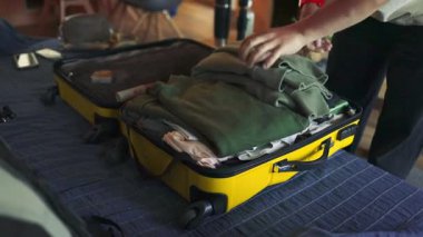 Bir insan düzgün katlanmış kıyafetlerini sarı sert kabuklu bir bavula koyar ve bir yolculuğa hazırlanır. Video, ev içi seyahat rutinini yakalıyor. Giysileri dikkatlice yerleştirirken ellerini gösteriyor.