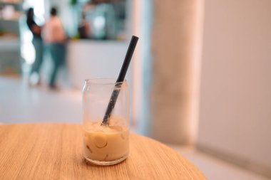Parlak, modern bir kafede, ahşap bir masanın üzerinde siyah pipetle kısmen bitmiş buzlu kahve bardağı. Arka planda tezgahın yanındaki insanlar ilgisiz ve rahat bir ortam yaratıyor.