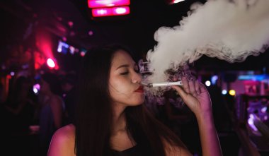 Kadın, gece kulübü barında elinde buhar cihazıyla otururken sigara içiyor. Sağlıksız davranış konsepti için elektronik sigaralı kadın.
