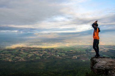 Adam yükselen bir uçurumun tepesinde duruyor, kolları zaferle yükseliyor, geniş bir tarımsal manzaraya bakıyor. Uçsuz bucaksız tarım arazileri aşağılara kadar uzanıyor, nefes kesici bir manzara sunuyor..