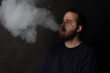 Sakallı adam siyah arka planda dururken büyük bir duman püskürtüyor.