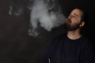 Sakallı adam siyah arka planda büyük bir duman bulutu salıyor.