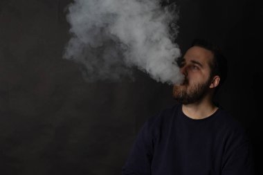 Sakallı genç adam karanlık ve kasvetli bir atmosferde büyük bir duman bulutu salıyor.