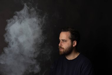 Siyah arka planda dumana bakan sakallı genç bir adam.