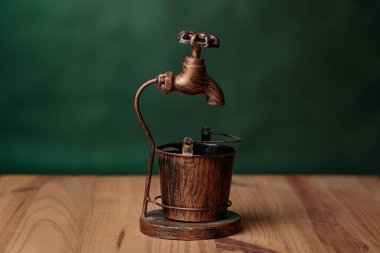 Antika bronz musluk, yeşil arka planda duran ahşap bir masaya yerleştirilmiş bir kovaya dökülüyor.