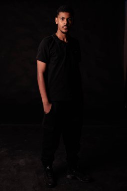 Stüdyo portresi, siyah elbiseli, koyu bir arka planda duran genç bir adam.