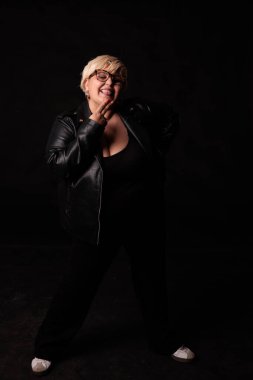 Siyah elbiseli, gözlüklü, gülümseyen ve stüdyoda siyah arka planda poz veren iri yarı bir kadın.