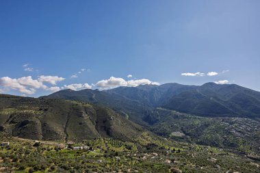 Sersemletici manzara dağların yeşil bir vadi üzerinde yükseldiğini gösteriyor. Evlerle benekli, açık mavi gökyüzünün altında az bulutlu.