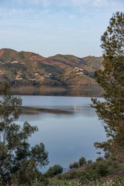 Guadalhorce rezervuarının sakin suları günbatımında tepeleri ve gökyüzünü yansıtıyor, Malaga, İspanya 'daki yemyeşil ağaçlar tarafından çerçevelenmiş,