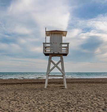 Boş beyaz cankurtaran kulesi kumlu bir sahilde duruyor bulutlu bir gökyüzünün altında sakin bir denize bakıyor.