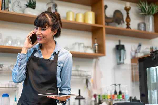 Latino female coffee shop owner or barista talking on the phone receiving customer order at the coffee shop. Smiling for successful business.