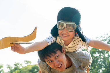 Asian boy wearing pilot helmet playing airplane toy on father back at park. dad support kid dream goal or aspiration. life living and family insurance concept. copy space