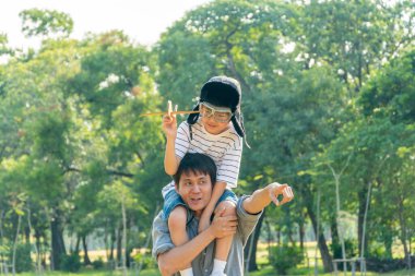 Asian boy wearing pilot helmet playing airplane toy sitting on father shoulder at park. dad support kid dream goal or aspiration. life living and family insurance concept