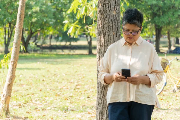 Asian senior woman standing at tree in the park playing on mobile phone. reading news, browsing the internet or playing game