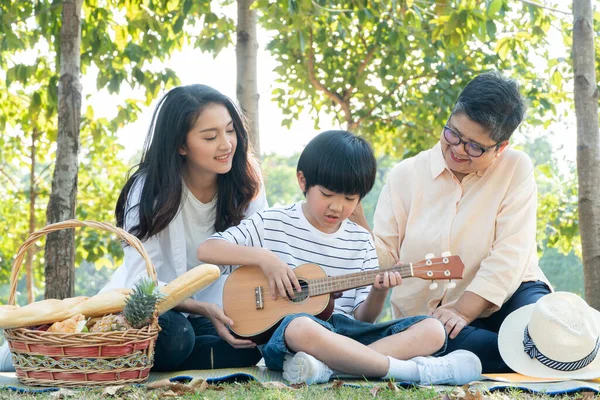 Happy Asian mother and grandmother watching son playing ukulele guitar while picnic at park, family having fun and spend time together on weekend, insurance concept of love and sharing