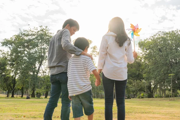 Back view of happy Asian family walking at park in summer season, mother holding son hand and colorful wind turbine, father touch his head with love, family activity lifestyle insurance concept