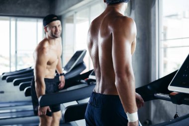 Two topless man exercise walking on treadmill and talking together. People workout lifestyle at fitness.