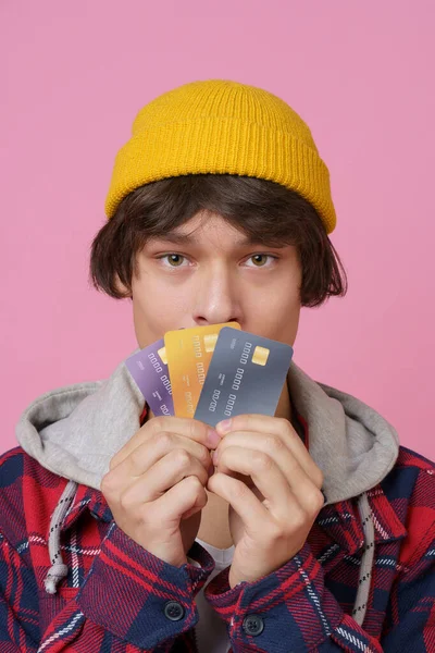 ハンサムな若いティーンエイジャーの男17年代はピンクの背景に隔離されたクレジット デビットカードの帽子を身に着けているし 再生ジャケットを保持します 銀行カードと現金を持つ若い男の肖像画 銀行の概念 — ストック写真