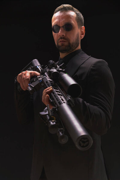 Secret service man in black suit, bodyguard or special forces agent man with gun isolated on black background. Man in black suit holding assault rifle. Spy, police man in black suit and sunglasses.