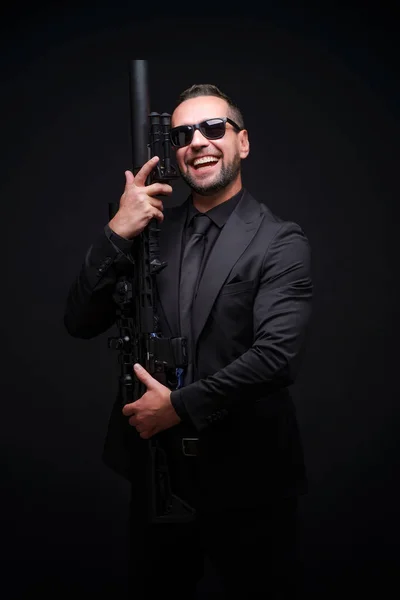 In love with his rifle man in black suit and sunglasses. Man holding assault rifle. Secret service man in black suit, bodyguard or special forces agent man with gun isolated on black background.