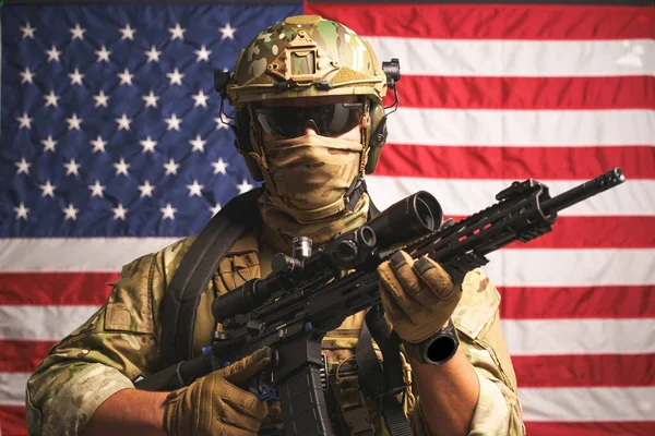 Army soldier in camouflage clothing holding riffle. Flag of United States of America on background. Soldier laid hands on machine gun dressed in ammunition posing on USA flag on background.
