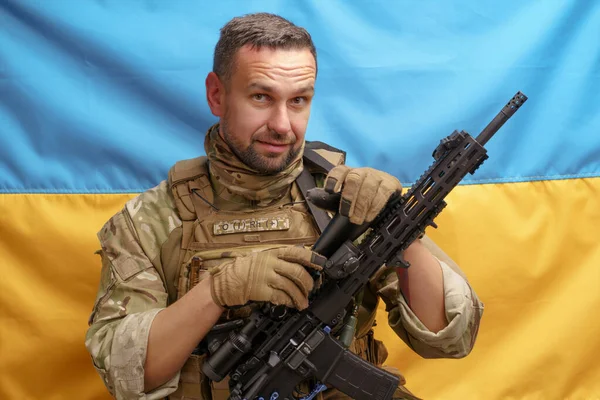 Soldier of Ukrainian Army handsome man holding rifle in a camouflage ammunition posing on Ukrainian flag background. Brave Ukrainian army man with weapon. The flag of Ukraine on background.