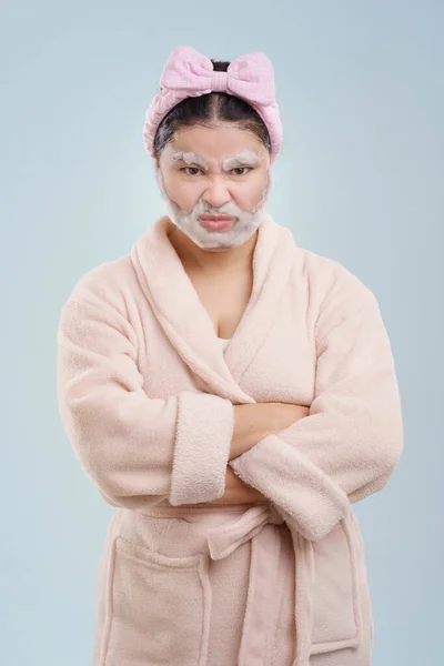 Mid aged Asian woman in bathrobe, with cosmetics foam shaped like beard on her face. She appears playful and humorous while engaging in her beauty routine, representing quirky and creative approach to