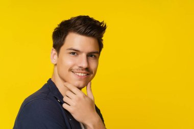 Elleri çenesine değen gülümseyen genç adamın yakın plan portresi güven ve pozitiflik havasını yansıtıyor. Görüntü onun gençlik güzelliğini ve benzersiz bireyselliğini vurguluyor.